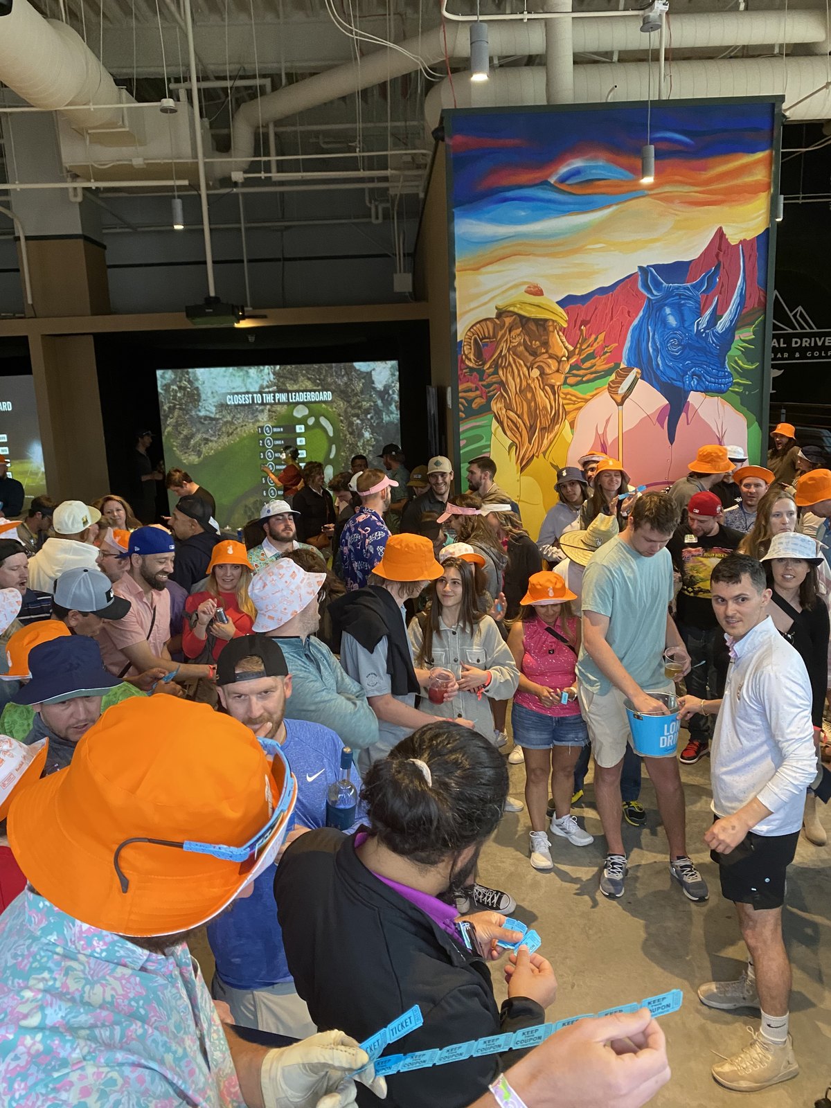 A crowd in bucket hats at a golf simulator night, with a closest-to-the-pin leaderboard on screen and raffle tickets in hand